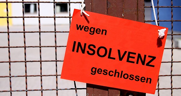 Een fel oranje bord met de tekst „wegen INSOLVENZ geschlossen“ hangt aan een hek. Het bord staat symbool voor een faillissement en insolventieprocedure in Duitsland. De afbeelding benadrukt de gevolgen van financiële problemen voor ondernemingen en illustreert het thema bedrijfsbeëindiging en insolventierecht.