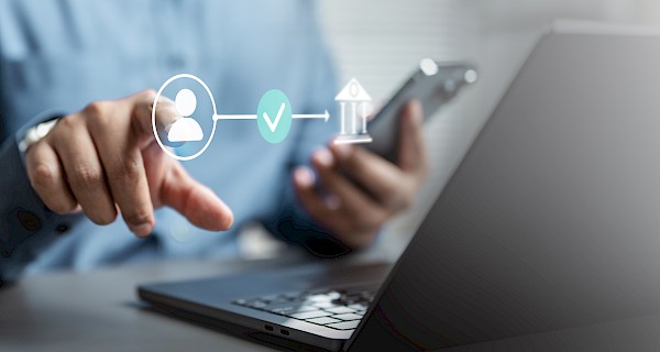 De foto toont een persoon die met één hand op een laptop werkt en met de andere hand een smartphone vasthoudt. Op de voorgrond is een digitale illustratie te zien met pictogrammen die een transactie of verificatie voorstellen: een gebruikersicoon, een vinkje en een bankgebouw. De afbeelding verwijst duidelijk naar een online betaling, digitale identificatie of beveiligde verificatieprocedure.